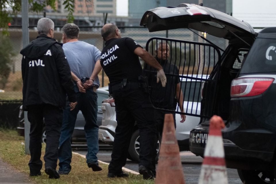 Homem de camisa cinza é retirado de viatura por policiais