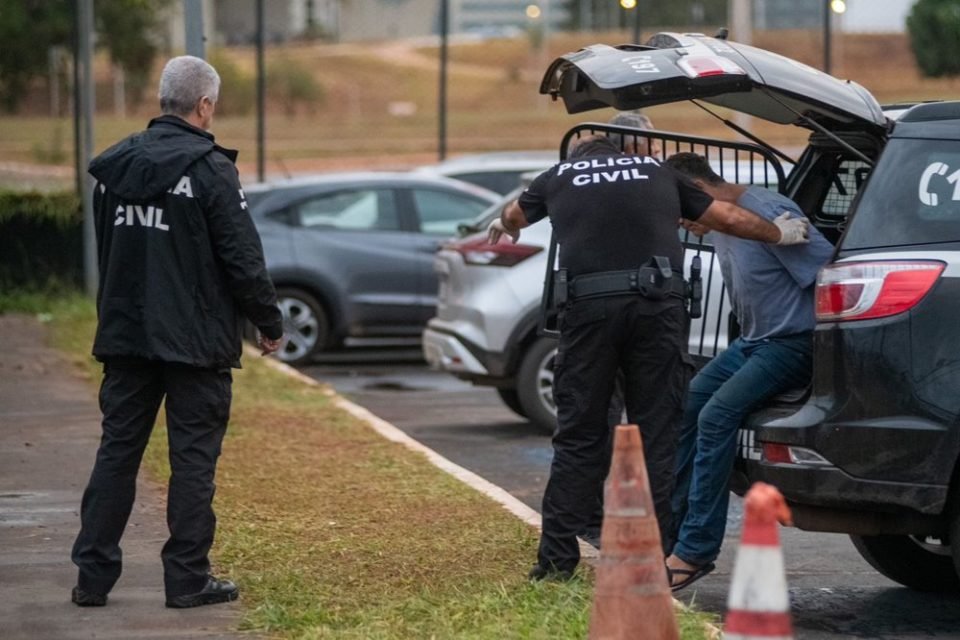 Homem de camisa cinza é retirado de viatura por policiais