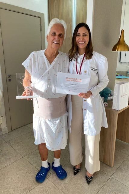 Foto mostra um senhor de cabeça branca e roupa de hospital de pé, segurando um diploma, com uma mulher de jaleco branco. Eles estão em um hospital - Metrópoles