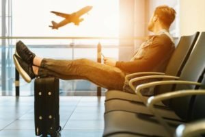 Fotografia colorida mostrando homem sentado em cadeira de aeroporto com pés sobre a mala e olhando para um avião decolando-Metrópoles