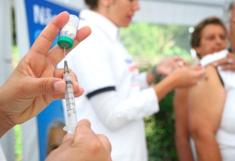 foto colorida, em primeiro plano enfermeiro segura seringa de vacina da gripe; um pouco adiante, enfermeiro com jaleco aplica vacina em pessoa - Metropoles