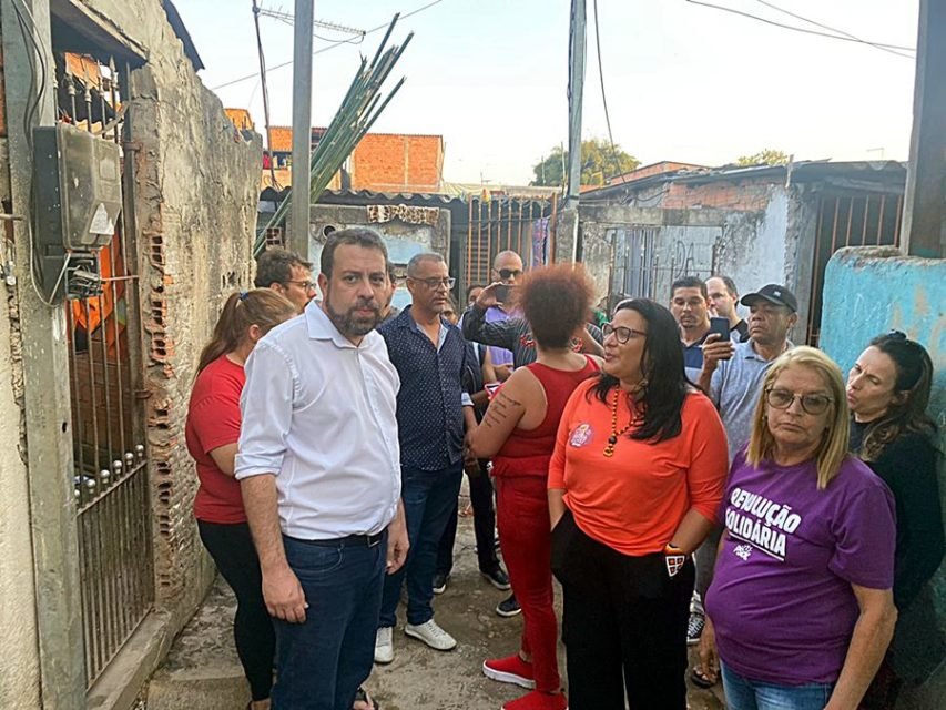 Imagem colorida mostra Guilherme Boulos homem branco de cabelo e barba castanhos, de camisa branca e calça jeans, em meio a um grupo de pessoas em uma viela da comunidade São João, em São Mateus, zona leste de São Paulo - Metrópoles