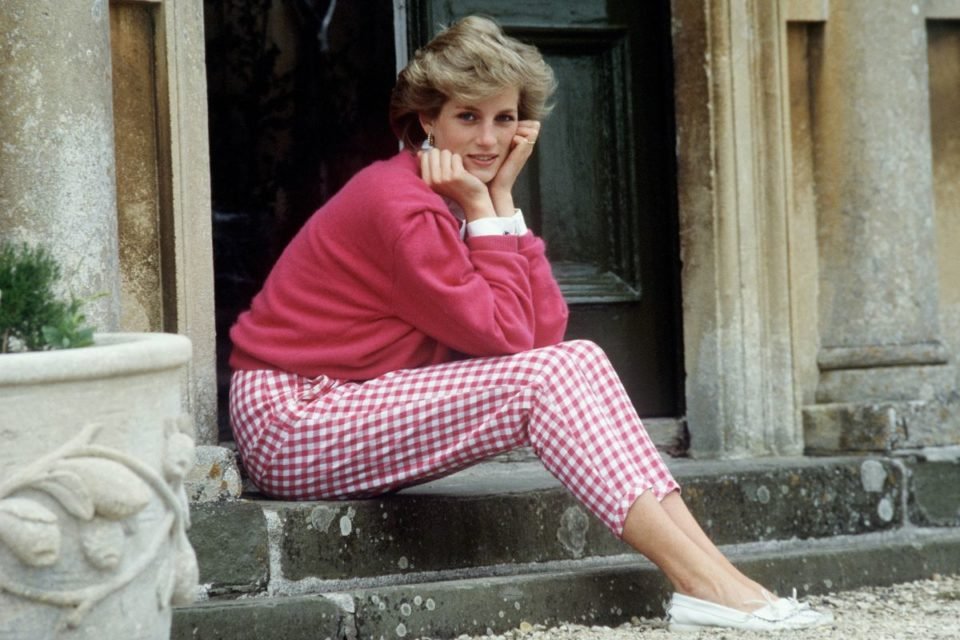 Princesa Diana sentada na calçada com um suéter cor-de- rosa e uma calça rosa claro. Ela está com as mãos no queixo olhando para a câmera - Metrópoles