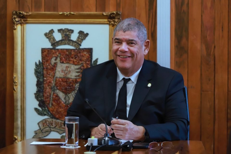 Imagem colorida mostra Milton Leite, homen negro, grisalho, de terno azulm, sentado em uma mesa de madeira em uma sala com paredes de madeira com um brasão da cidade de São Paulo atrás dele. Leite sorri na foto - Metrópoles