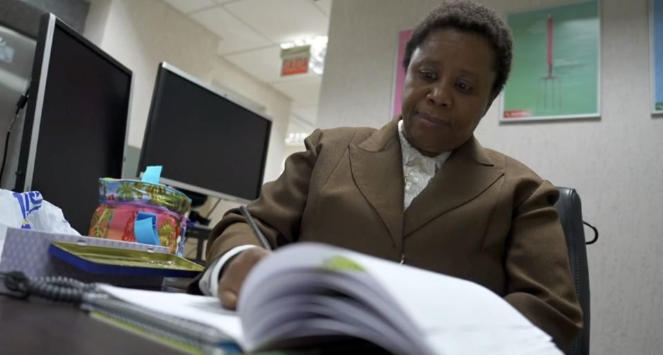 Imagem colorida mostra Sonia Maria de Jesus, uma mulher negra, com cabelos amarrados, usando um terno marrom e uma camisa branca, escrevendo algo em um caderno - Metrópoles