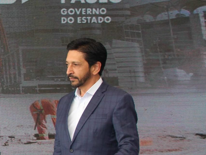 Imagem mostra Ricardo Nunes de terno azul e camisa azul, olhando para sua direita, na frente de um banner do governo do estado, da cintura para cima - Metrópoles