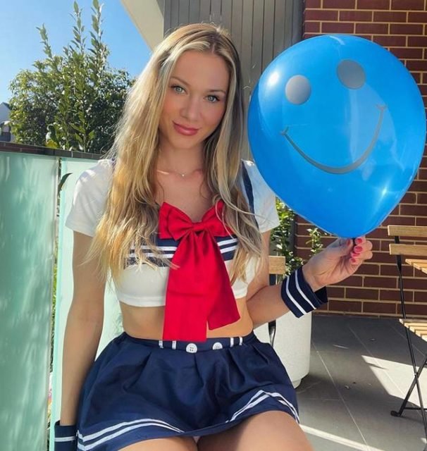 Foto colorida de uma mulher ajoelhada com uma fantasia de marinheira e segurando um bal&atilde;o azul com um desenho de sorriso - Metr&oacute;poles