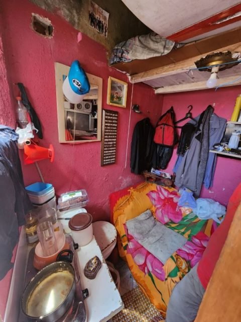 Imagem colorida mostra casa com parede rosa, roupas penduradas em cabides na parede, sofá com capa colorida e prateleira com restos de comida. Metrópoles