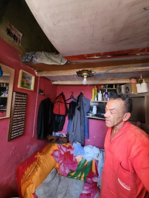 Imagem colorida mostra homem com blusa vermelha em casa com parede rosa, roupas penduradas em cabides na parede, sofá com capa colorida. Metrópoles