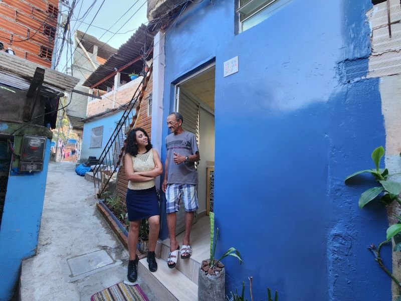 imagem colorida mostra mulher de cabelos cacheados de braços cruzados enquanto olha para homem. o homem está em frente a porta de uma casa, veste camiseta cinza e shorts claro. a casa tem parede azul. metrópoles