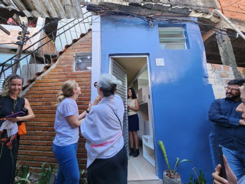 Imagem colorida mostra casa de 4 metros quadrados em São Paulo. As paredes são azuis, tem duas pessoas do lado de fora olhando e uma na porta do lado de dentro - Metrópoles