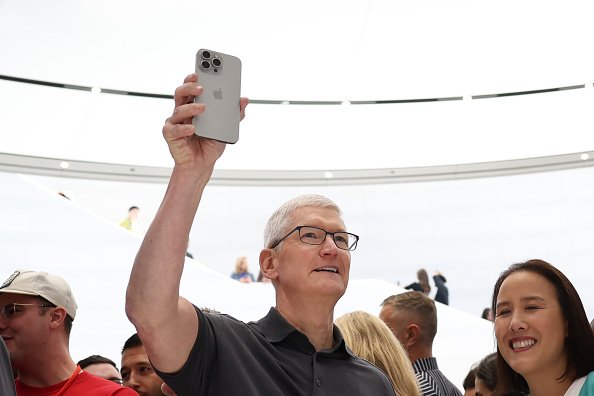 Tim Cook, CEO da Apple, levanta o braço segurando um iPhone 15. Ele usa óculos, tem cabelos grisalhos e veste uma camisa escura - Metrópoles