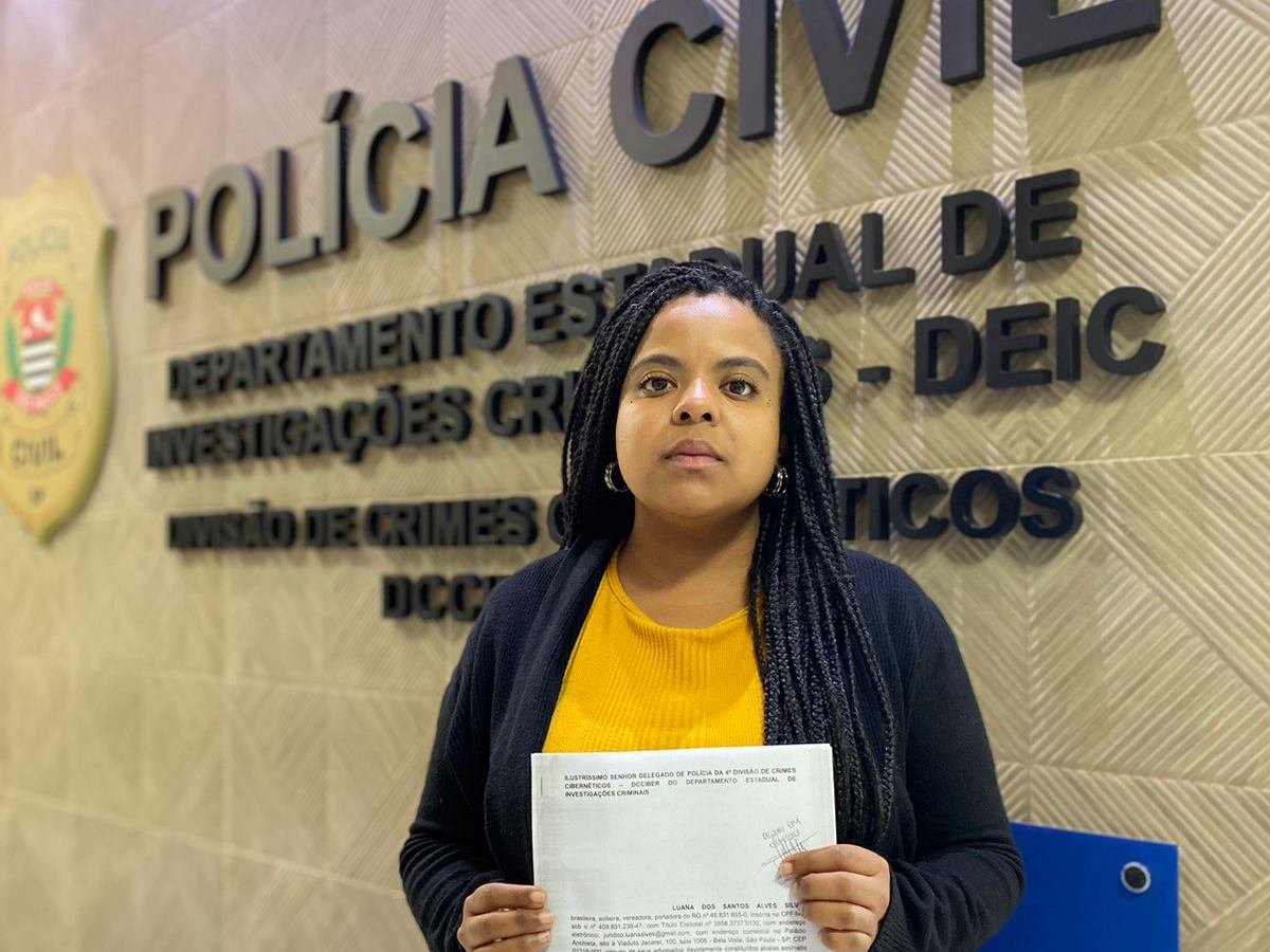 Imagem colorida mostra a vereadora Luana Alves, mulher negra de cabelos pretos, blusa preta e camisa amarela, na frente de uma parede com letreiro da polícia - Metrópoles