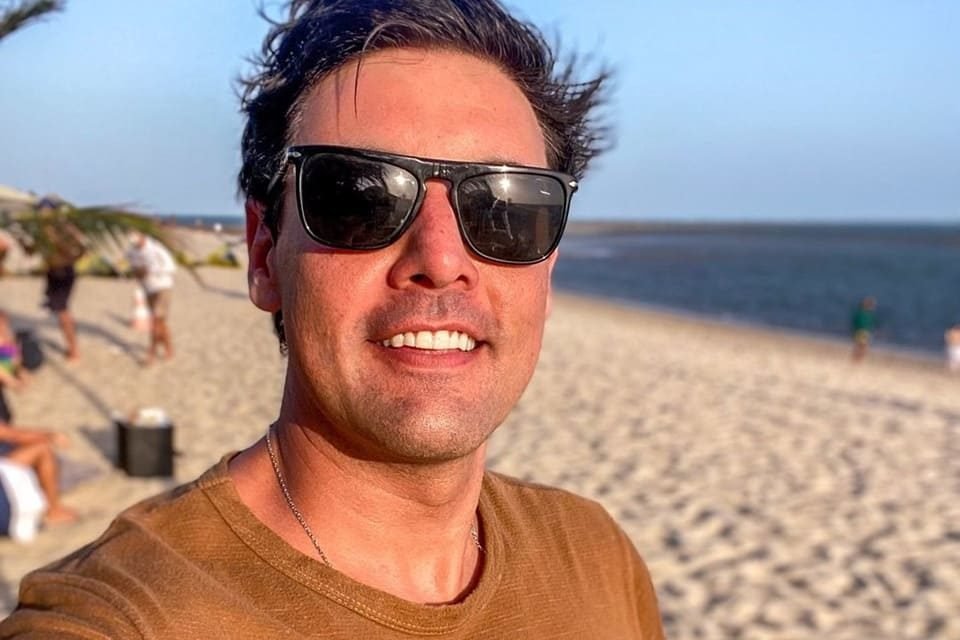 Bruno De Luca posa de óculos escuros e blusa marrom na praia - Metrópoles