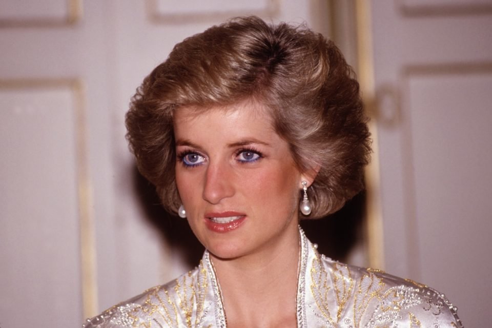 Foto colorida. Princesa Diana, uma mulher branca, loira e de olhos azuis olha fixamente para a frente. Ela usava roupas claras - Metrópoles