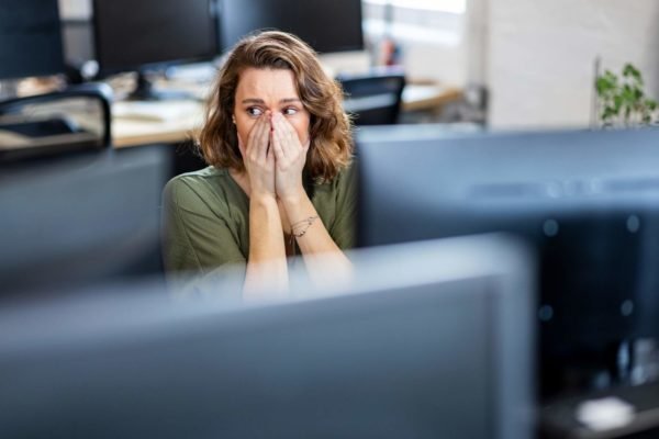 Foto mostra mulher angustiada com as mãos no rosto ao lado de vários monitores de computador