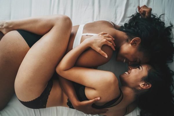 Foto colorida de duas mulheres deitadas na cama de lingerie abraçadas olhando uma para a outra - metrópoles