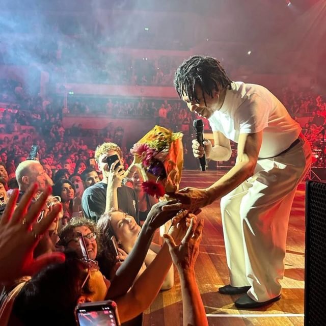 Foto colorida de Djavan no palco com a plateia - Metrópoles