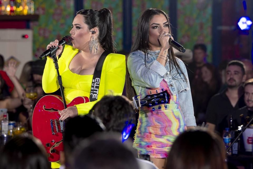 foto colorida da dupla Day e Lara, uma delas de vestido amarelo fluorescente e a outra de vestido estampado com jaqueta jeans, em cima de palco cantando - metrópoles