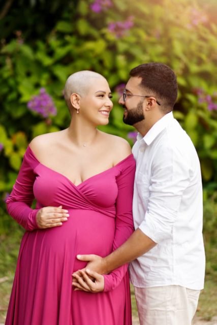 Casal posando para foto. Mulher de cabeça raspada e vestido rosa e homem segurando a barriga dela - Metrópoles