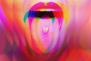 Foto colorida e com efeito psicodélico de uma mulher com a língua para fora com uma pílula em formato de coração - Metrópoles