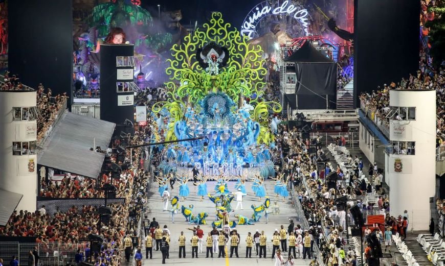 Imagem colorida mostra um dos desfiles de escola de samba em São Paulo, com alas em primeiro plano e um carro alegórico atrás - Metrópoles