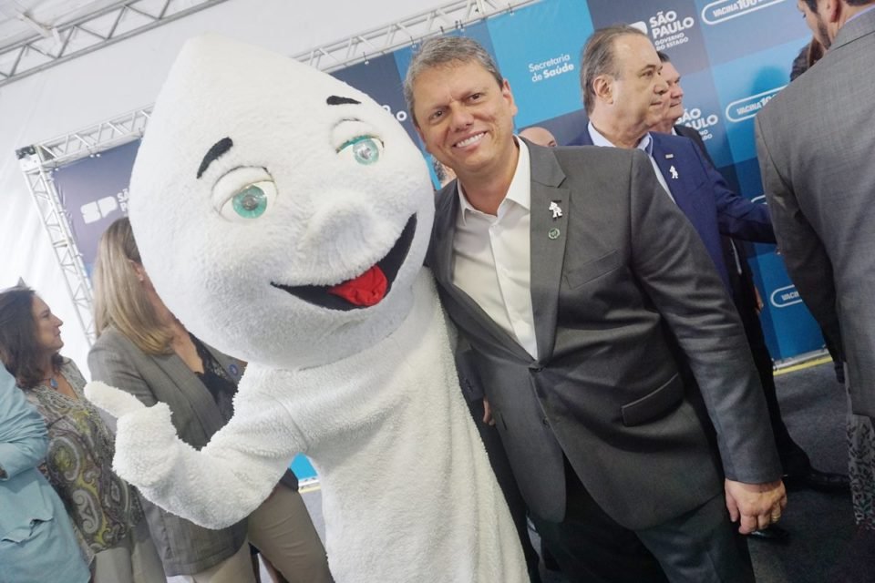 Imagem colorida mostra homem fantasiado com a roupa do "Zé Gotinha" abraçado com o governador Tarcísio de Freitas, homem branco, grisalho, de terno cinza, em um evento sobre vacinação - Metrópoles