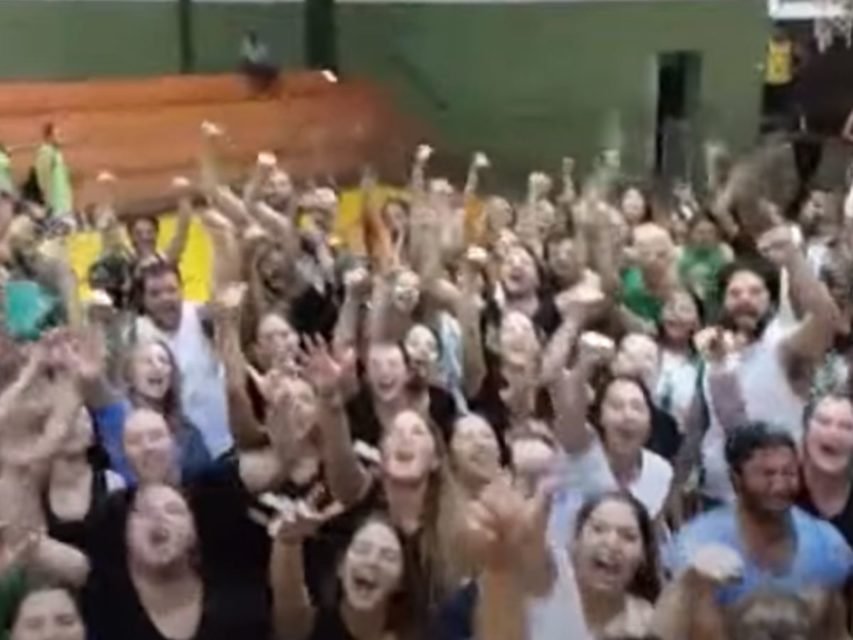 Imagem colorida mostra estudantes cantando hino de faculdade