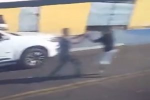 Imagem colorida mostra dois homens brigando na frente de um carro branco. O idoso de calça comprida acabou levando um tiro do jovem de bermuda - Metrópoles