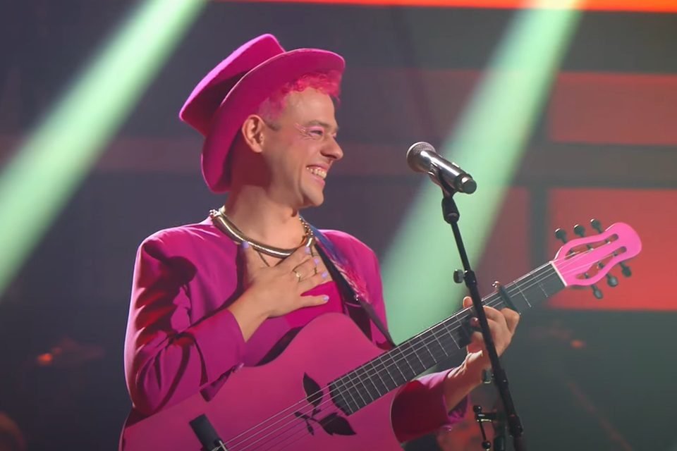 Homem branco sorrindo em um palco. Ele usa um chapéu rosa, tem cabelo rosa, usa um blazer e uma blusa rosa e segura um violão rosa - Metrópoles