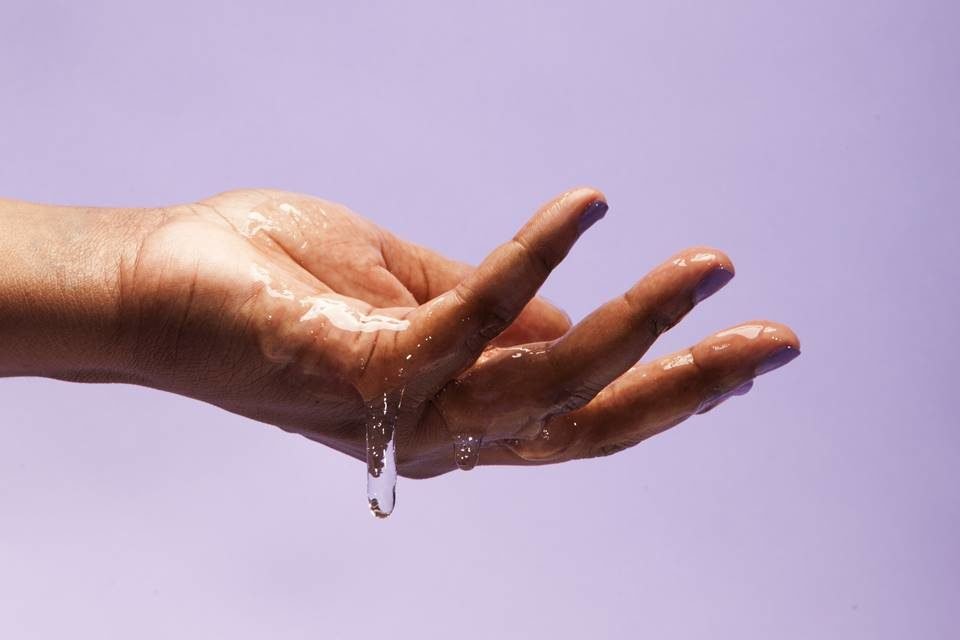 Foto colorida da mão de uma mulher cheia de um líquido transparente viscoso escorrendo em um fundo lilás - Metrópoles