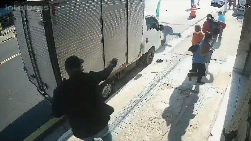 Imagem colorida mostra um bandido de blusa preta atirando em um PM à paisana que está correndo para se esconder na frente de um caminhão estacionado. Ele é observado por outro bandido de blusa vermelha - Metrópoles
