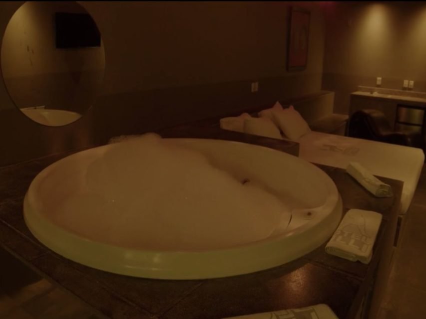 Foto colorida mostra banheira com espuma ao lado de cama de casal. Ambiente é escuro.