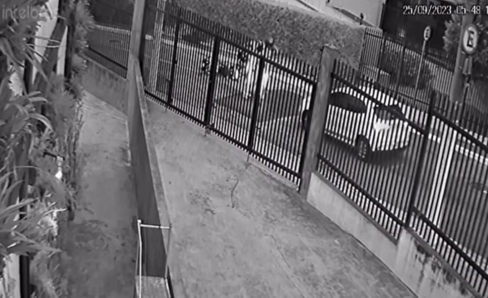 Imagem preta e branca com visão de dentro de portão para a rua; três homens estão na rua e um carro branco - Metrópoles