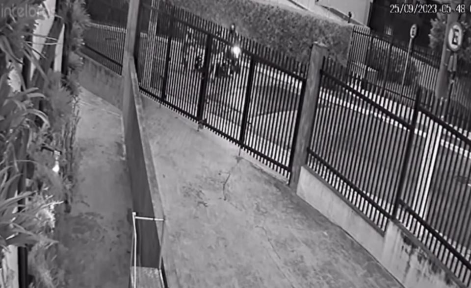 Imagem preta e branca com visão de dentro de portão para a rua; três homens estão na rua - Metrópoles