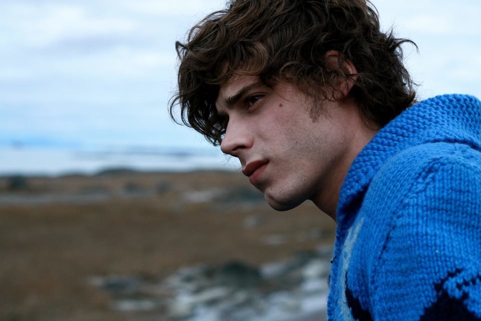 Foto colorida de Xavier Dolan. Ele é branco, usa moletom azul, tem cabelo preto e grande e está em uma praia - Metrópoles