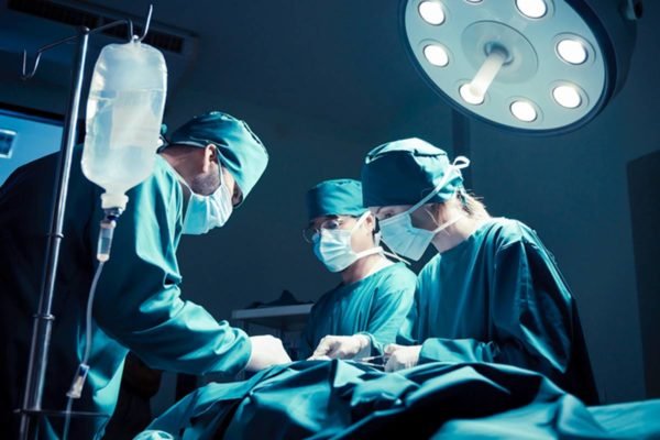 Imagem colorida de cirurgiões reundidos envolta de mesa de operação - Metrópoles