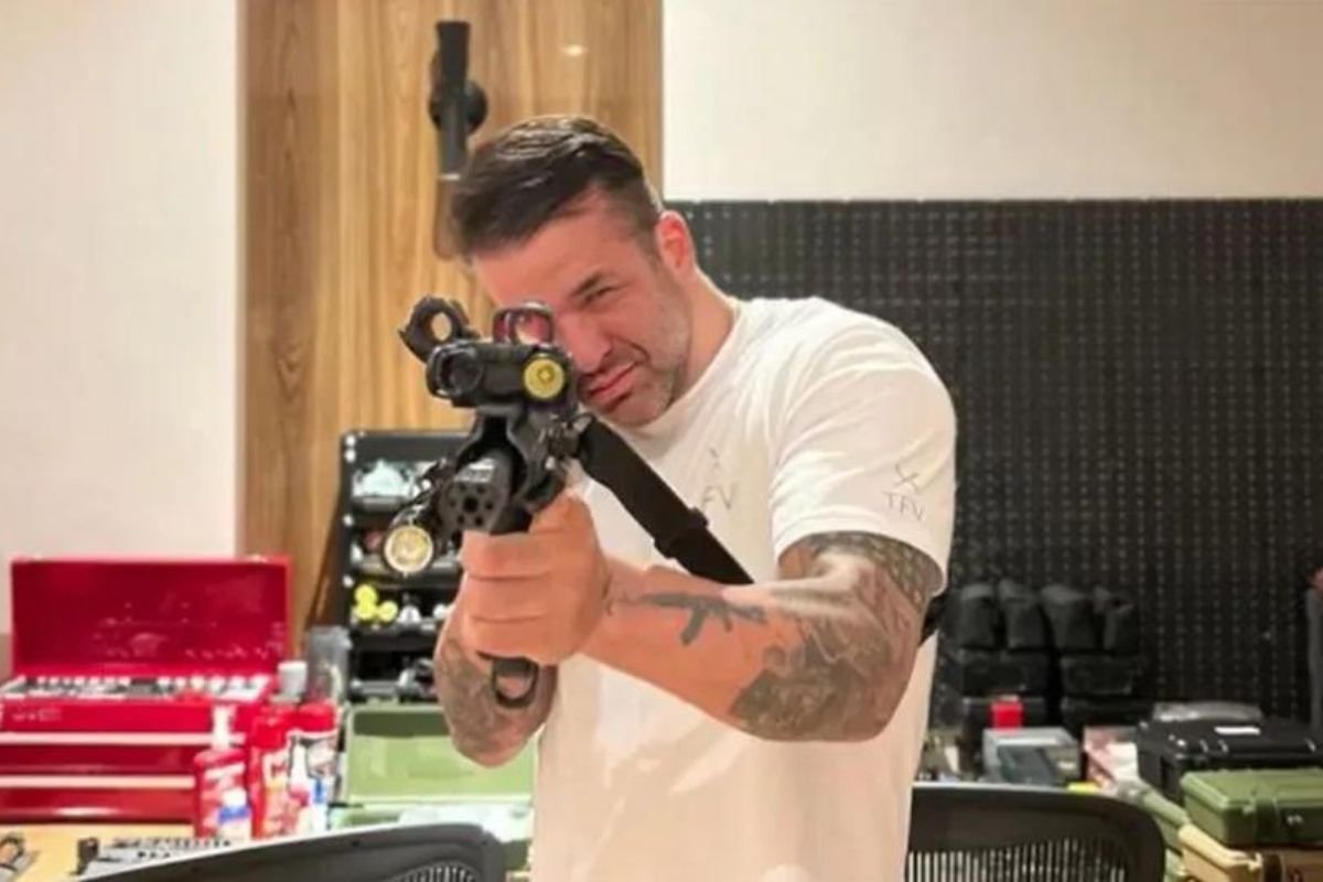 Imagem colorida de Thiago Brennand segurando arma de fogo. Ele é um homem branco, de camiseta branca e tatuagem no braço, e aponta a arma em direção da câmera - Metrópoles