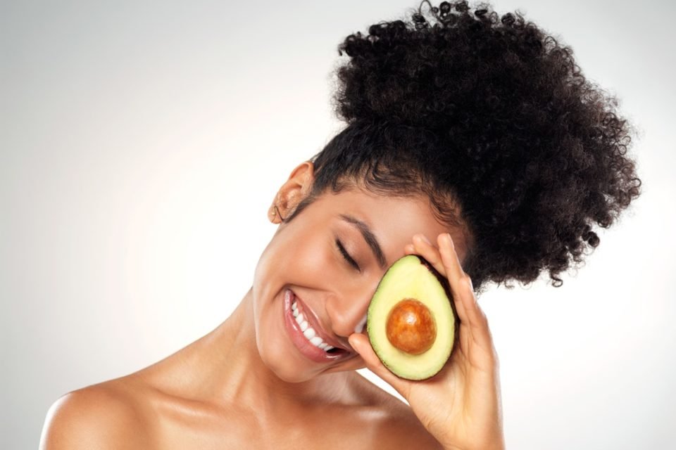 Foto colorida de mulher negra tampando o uma parte do rosto com a metade de um abacate - Metrópoles