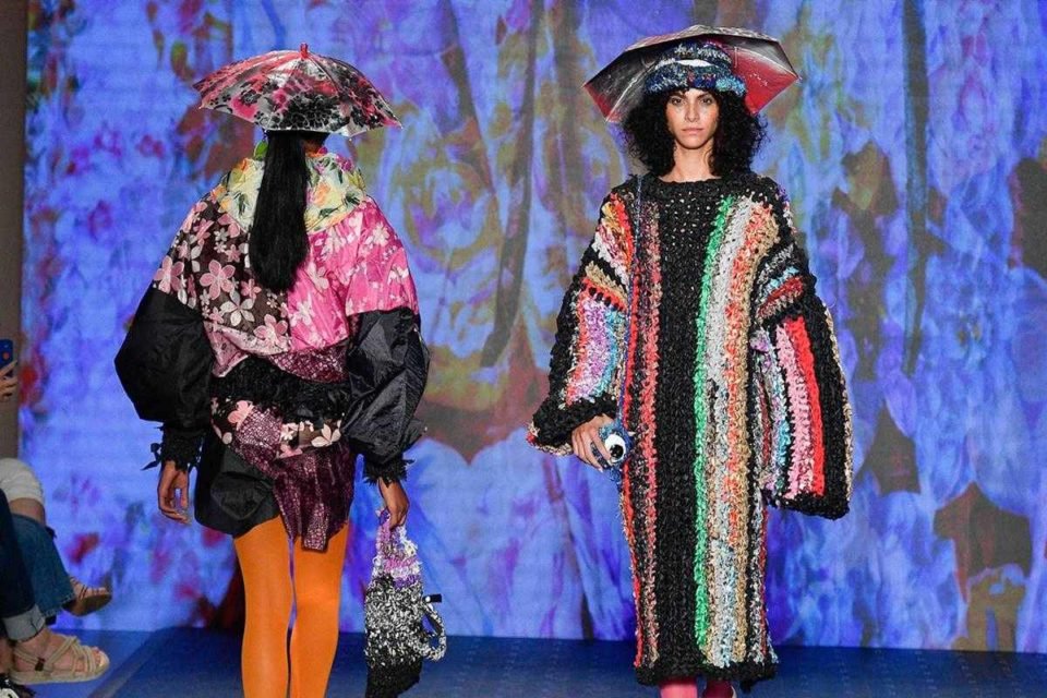 Em desfile de moda com fundo azul, duas modelos usam roupas coloridas com guarda-chuvas na cabeça - Metrópoles