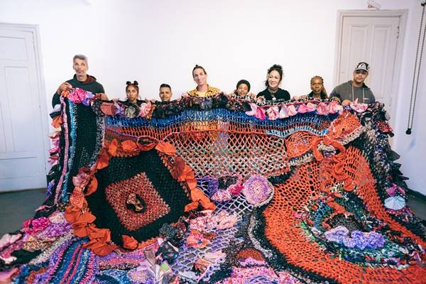 Peça colorida de tapeçaria segurada por um grupo de pessoas - Metrópoles