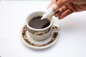 Imagem colorida. Imagem mostra uma xicara de café sobre um fundo branco e uma mão aplicando o adoçante - metrópoles