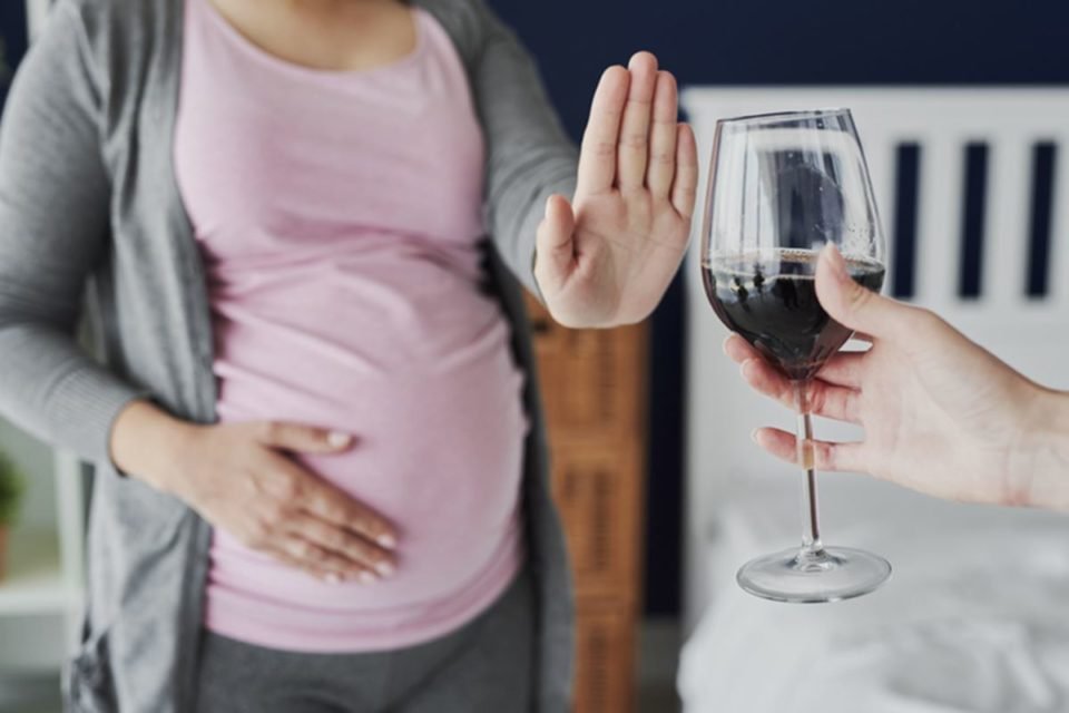 Foto colorida de mulher grávida com uma mão na barriga e a outra empurrando taça de vinho consumo de álcool na gravidez - Metrópoles