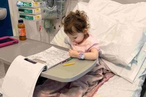 Foto mostra Grace Tudor pintando um desenho em uma cama de hospital após o diagnóstico do câncer