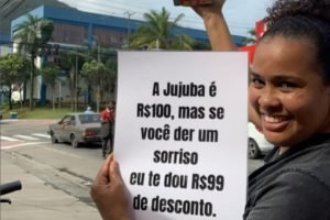 foto colorida da ambulante Bruna Correia, com cartaz em que anuncia que dá descontos em seus doces para quem sorrir - Metrópoles