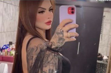 Imagem colorida mostra Paola Gobel, uma mulher de cabelos ruivos, branca, com tatuagens no braço direito, segurando um celular e tirando uma foto de lado diante de um espelho - Metrópoles