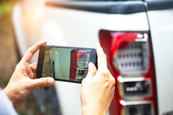 Imagem de homem tirando foto, com o celular, de carro batido, para acionar o seguro - Metrópoles