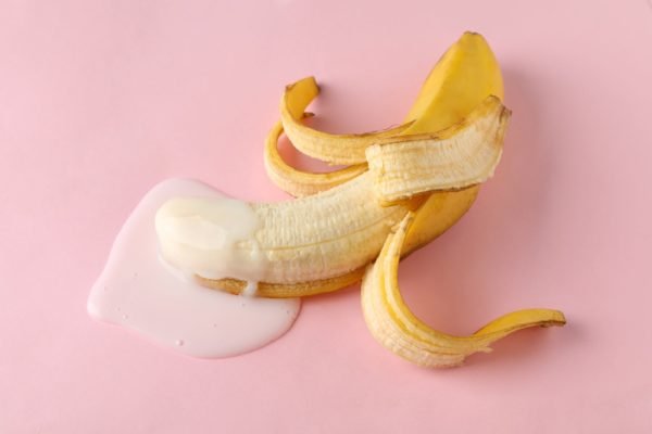 Foto ilustrativa em fundo rosa mostra banana descascada coberta com leite condensado, ilustrando o ato de ejacular - Metrópoles
