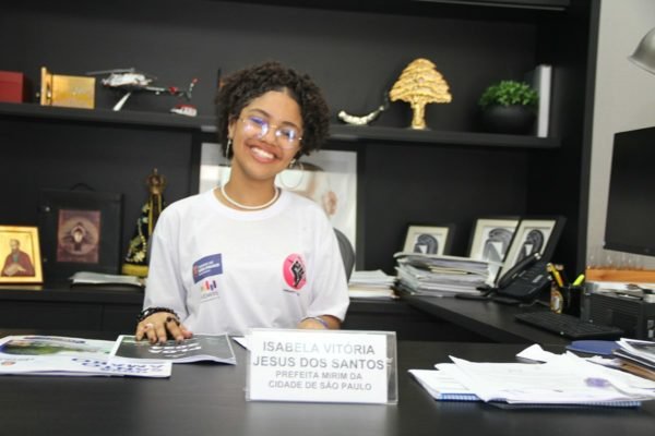 Imagem mostra a jovem Isabela dos Santos na mesa do prefeito de São Paulo. Ela, que foi eleita prefeita-mirim da cidade, é uma adolescente negra, de cabelo curto, usando óculos de grau e vestindo uma camiseta branca - Metrópoles