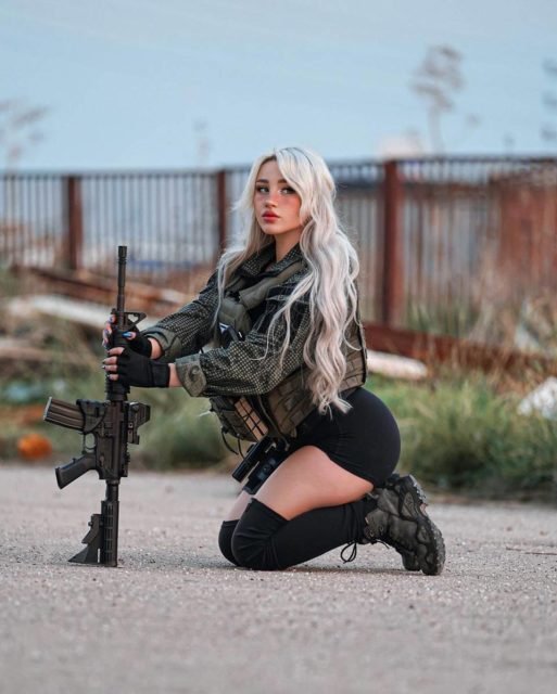 foto colorida de mulher de jqueta do exercito com short e meião pretos segurando arma de joelhos no chão - metrópoles